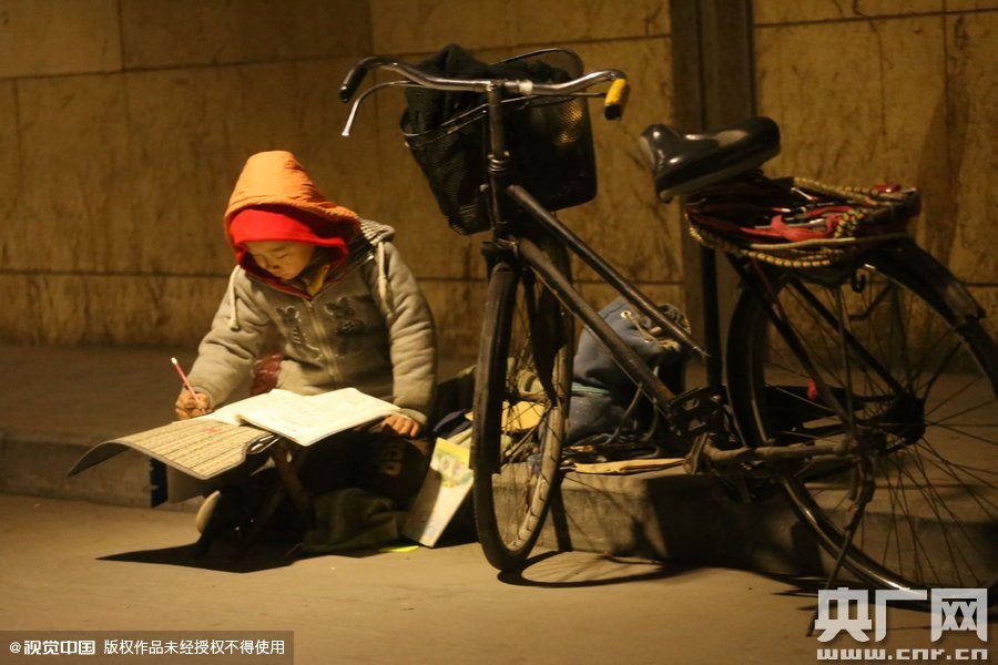 鄭州中原路京廣鐵路大橋下涵洞的馬路牙子邊，一位戴著紅色辣椒帽的小男孩趴在一個小馬扎上，上面鋪著一張車墊，在微弱的燈光下寫作業(yè)